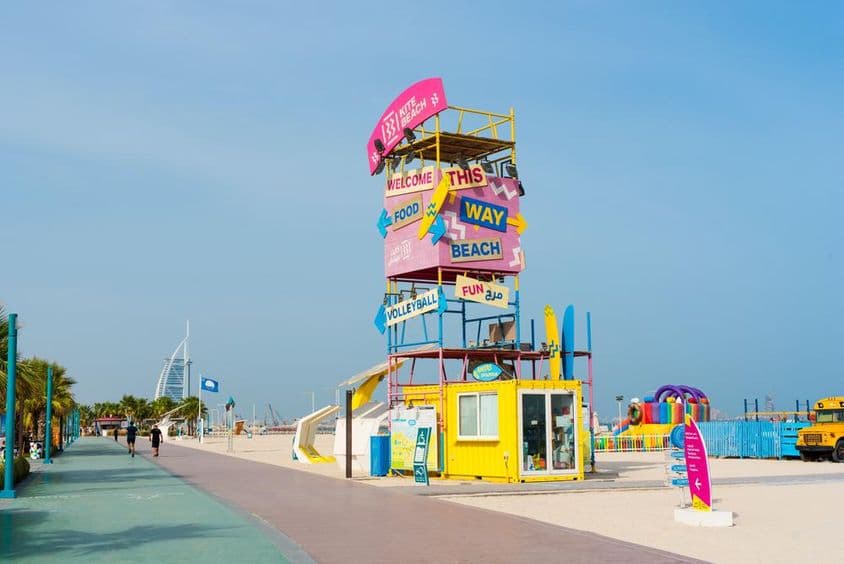 Kite Beach, Дубай: структура доброжелательства, знаки, Бурдж Аль Араб на заднем плане, беговая дорожка и песчаный пляж на переднем плане.