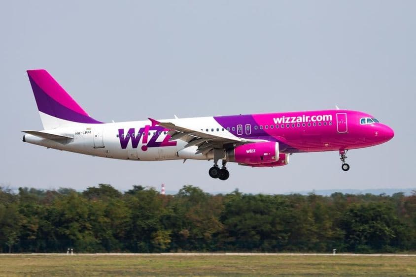 Самолет Wizz Air летит на низкой высоте с лесом на заднем плане.
