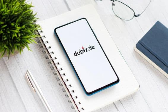 Телефон с запущенным Dubizzle на столе, под блокнотом, рядом с очками и небольшим зеленым растением.