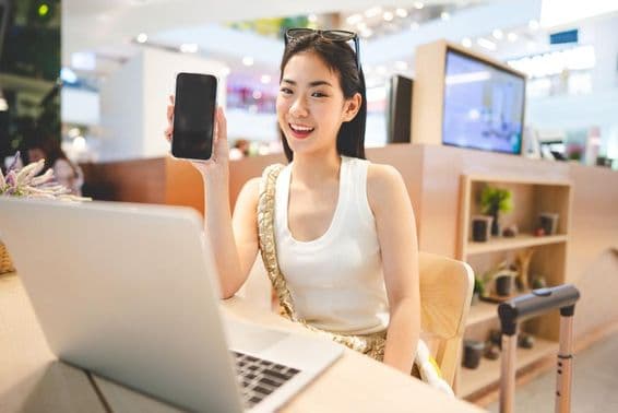 Девушка поколения Z работает, держит телефон, с ноутбуком перед ней и ручкой от чемодана рядом.