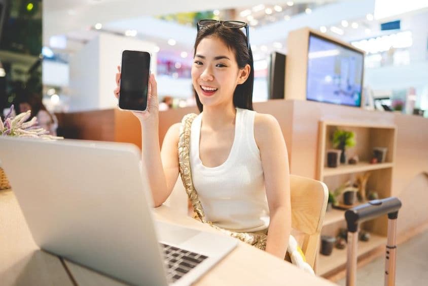 Девушка поколения Z работает, держит телефон, с ноутбуком перед ней и ручкой от чемодана рядом.