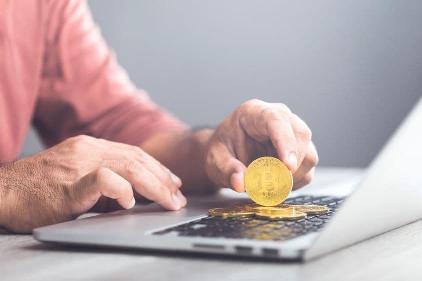 Мужчина держит золотую монету биткойна, используя ноутбук.