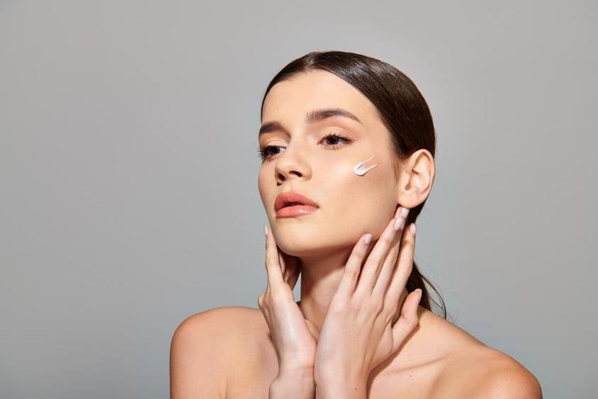 Молодая женщина с каштановыми волосами наносит крем для лица, излучая ощущение заботы о себе.