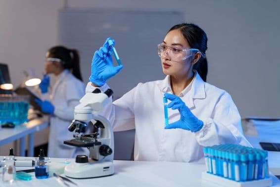 Азиатская женщина-учёный в лабораторном халате и перчатках сравнивает две пробирки.