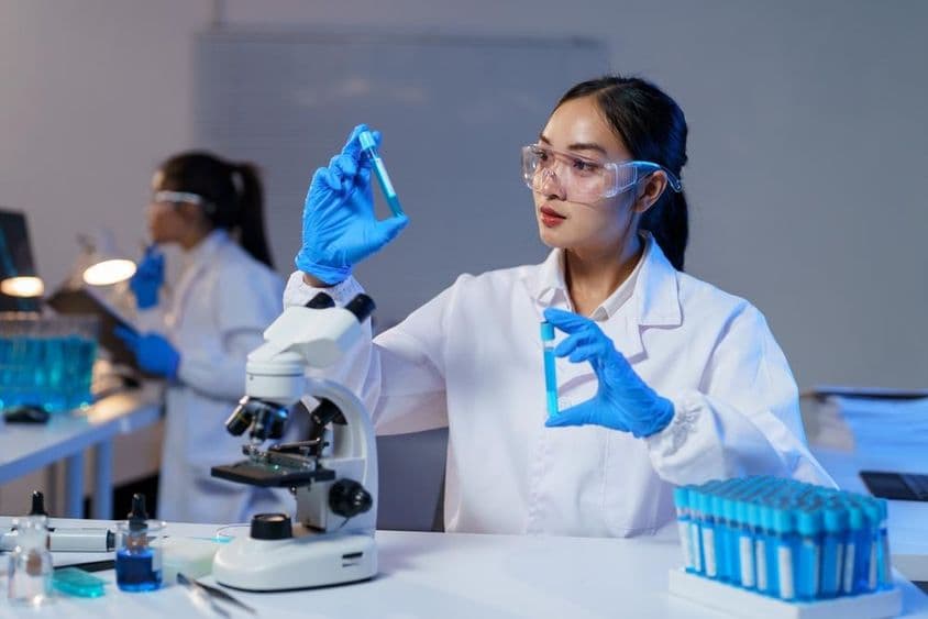 Азиатская женщина-учёный в лабораторном халате и перчатках сравнивает две пробирки.